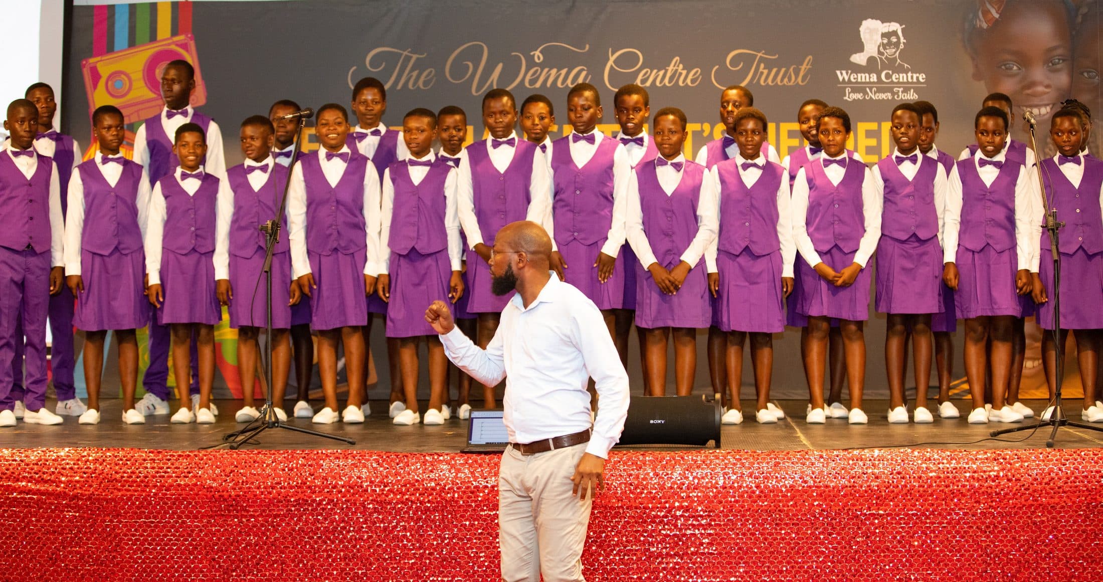 The Wema Centre Trust choir performing on stage in purple uniforms, led by their conductor